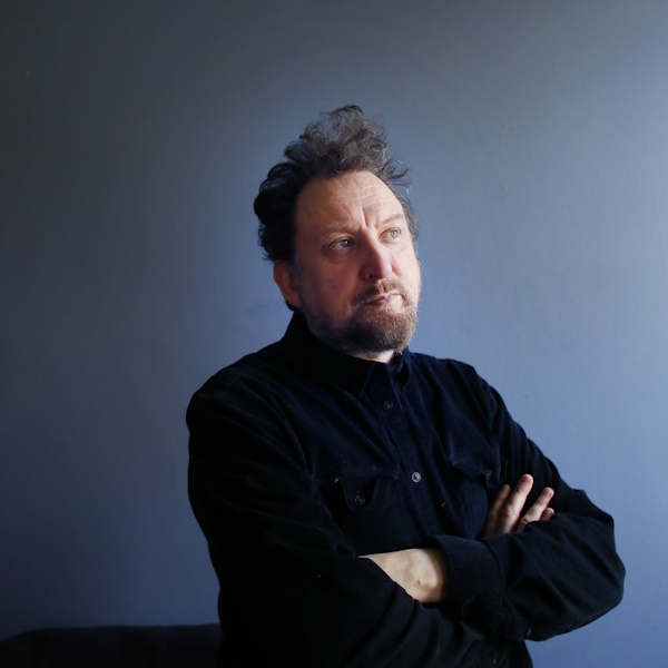 author Ewan Morrison dark clothes blue background looking pensive and upward, away from the viewer
