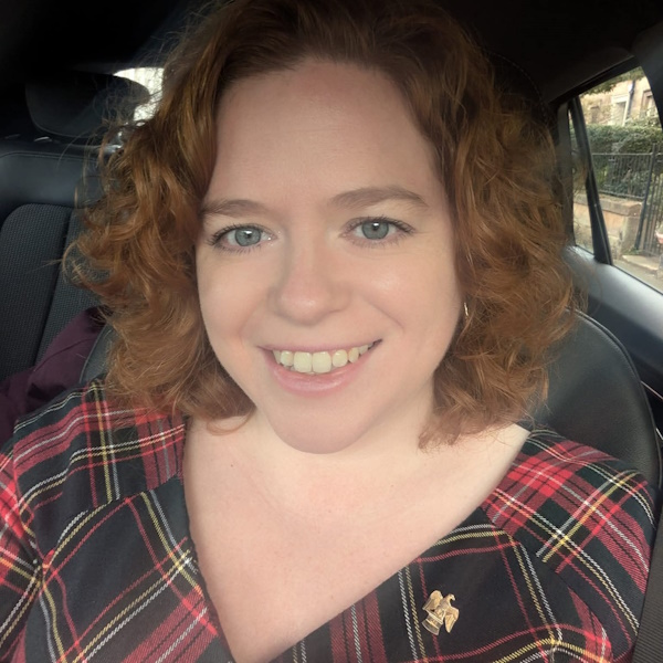 head and shoulders photograph selfie style photo of the Scottish Gaelic singer and poet Ceitidh Chaiumbeul, she is a smiling women under 40 with wavy red hair and wears a bold tartan dress
