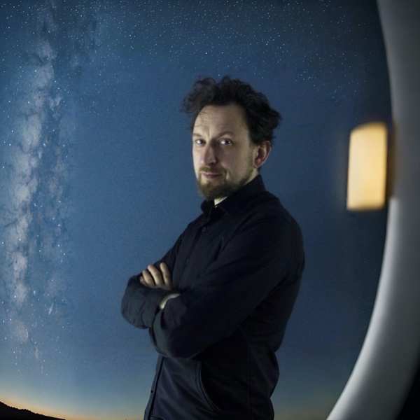 author ewan morrison is a man in a  dark suit with a neat beard and tousled hair and he stands before a view of the upper atmosphere with a minute part of a horizon slanted in the left foreground 