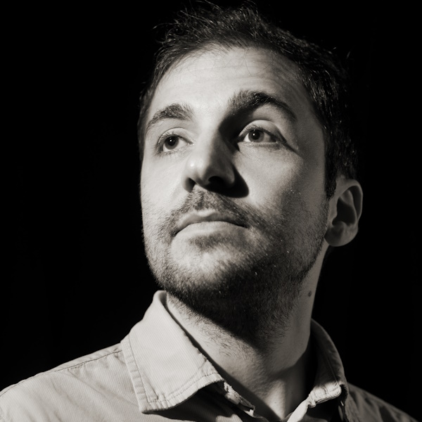 author Luca Serra pictured in black and white, he is a handsome man with a faint= beard and looks upward and into the distance behind the viewer